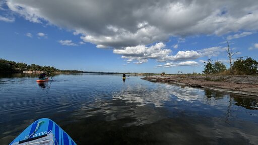 Photo of a chanel between two islands with a cloudy sky reflecting of the water, and a couple of paddleboarders heading through.