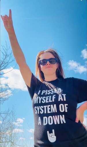 Woman making the sign of horns with her hand, wearing a black t-shirt that says "I pissed myself at System of a Down".